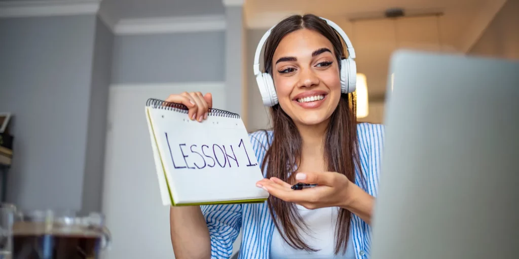 Online tutor wearing headphones and holding up a notebook that says, "Lesson 1."