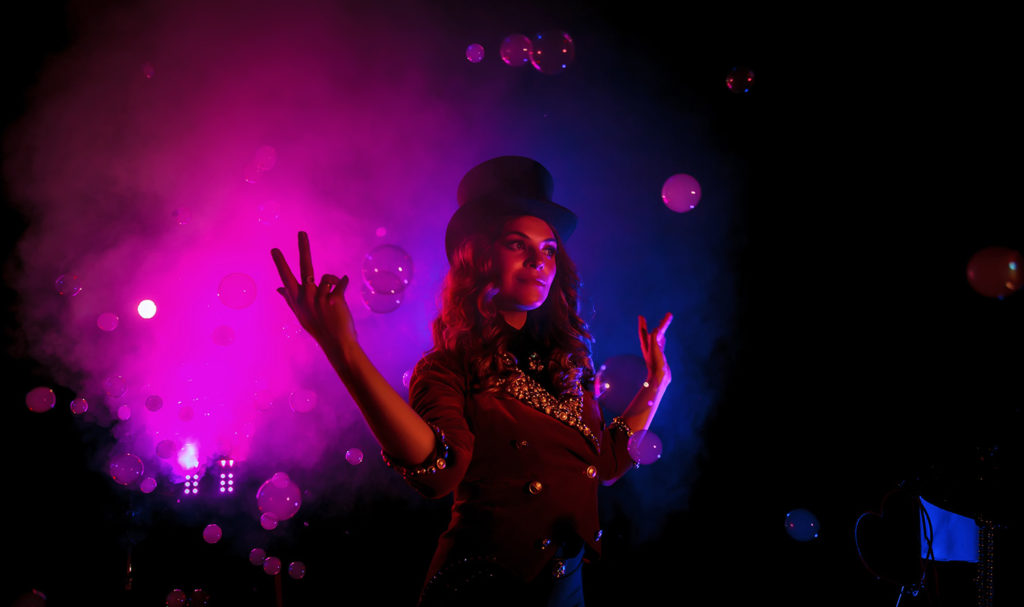 A magician insurance policyholder is illuminated on stage by pink and blue lights as bubbles and a fog machine surround her for her next trick.