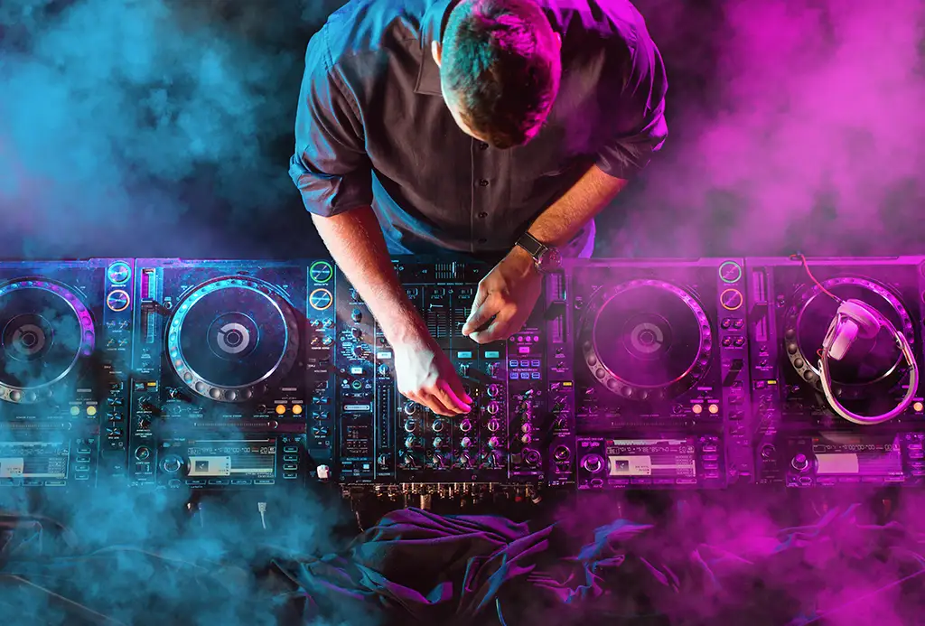 An overhead shot of a DJ at a turntable lit by purple and blue lights and surrounded by fog.