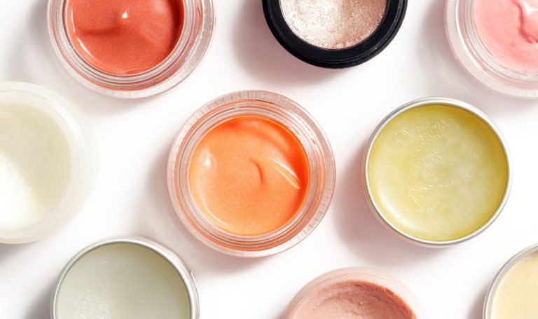 A variety of colorful beauty and skincare products sit in jars on a white surface and are viewed from above.