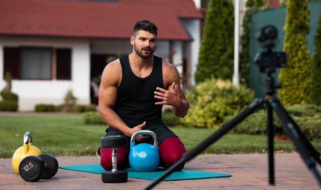 An online personal training is set up with weight and a a mat outside as he films himself demonstrating different workouts with a camera on a tripod.