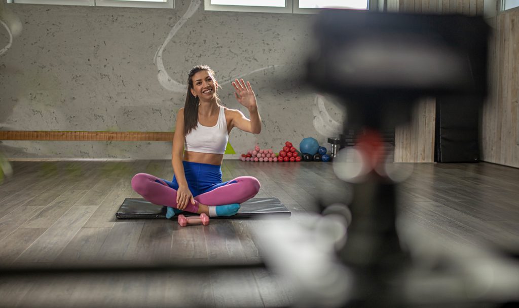 A young woman waves to her camera from her private studio where she films an online personal training video.