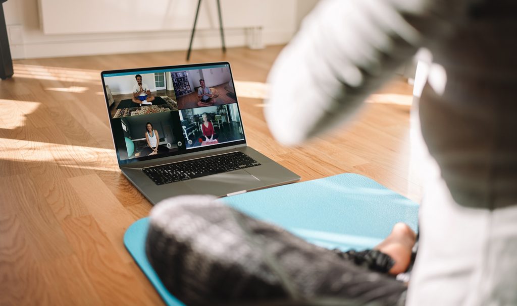 Someone sits on a light blue workout mat on the floor of their home as they watch a livestream fitness class on their computer with other virtual clients.