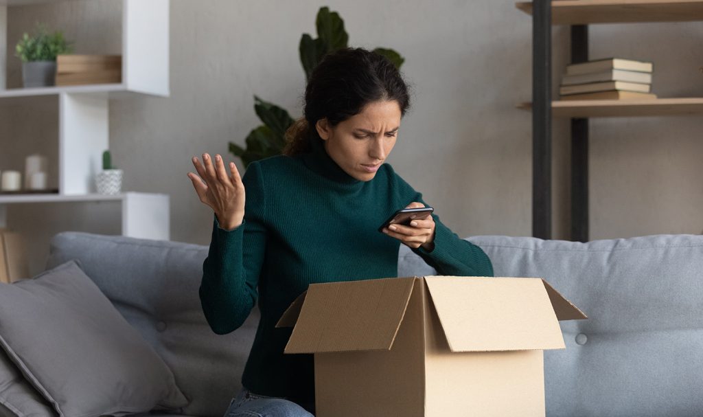 A woman sits on her couch upset as she films the inside of a box on her lap. A product arrived broken and she is documenting how it looked.