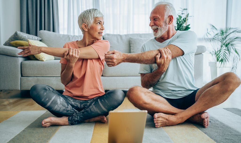 An older couple sits cross legged on yoga mats in their living room. They are stretching while following along with a video on an iPad propped up on the floor in front of them. These online personal training clients are discussing feedback they have on this program.