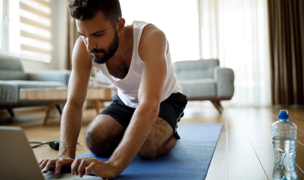 A virtual fitness instructor kneels on a blue yoga mat in his home as he types on his laptop and works on creating his online personal training packages.