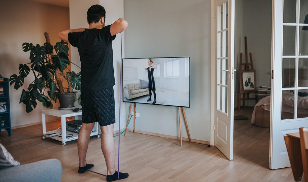 You are facing the back of a young man who is working out in his home with a resistance band and following an online personal training video on his television in front of him.