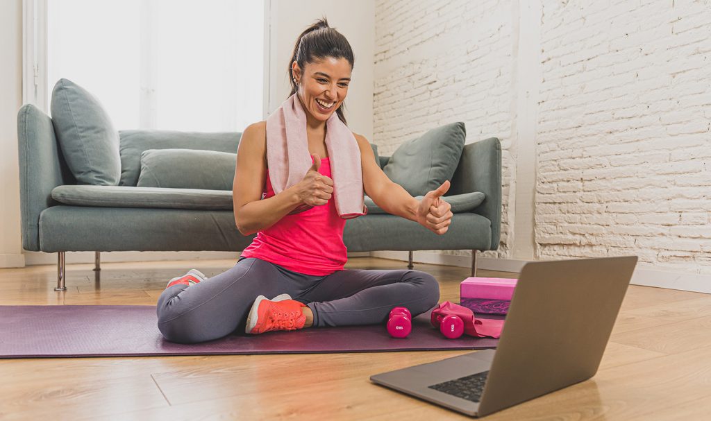 An online fitness instructor sits on a yoga mat in her home. She has pink weights next to her and a towel around her neck. She gives a thumbs up toward her laptop next to her as she cheers on a client she just trained with virtually.