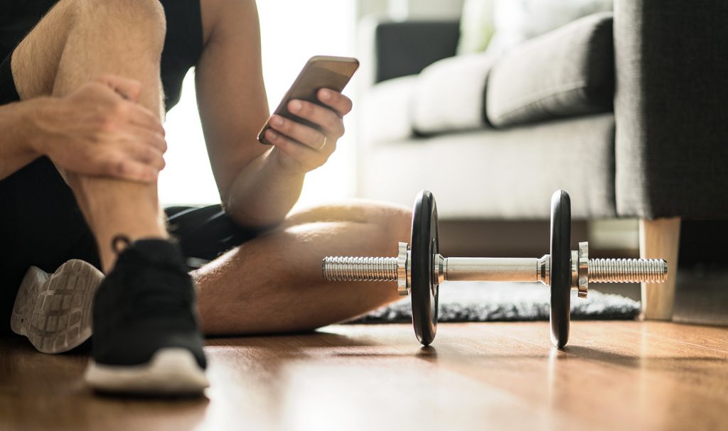 A virtual personal trainer sits on the floor of his home next to a weight as he texts a client during a workout.