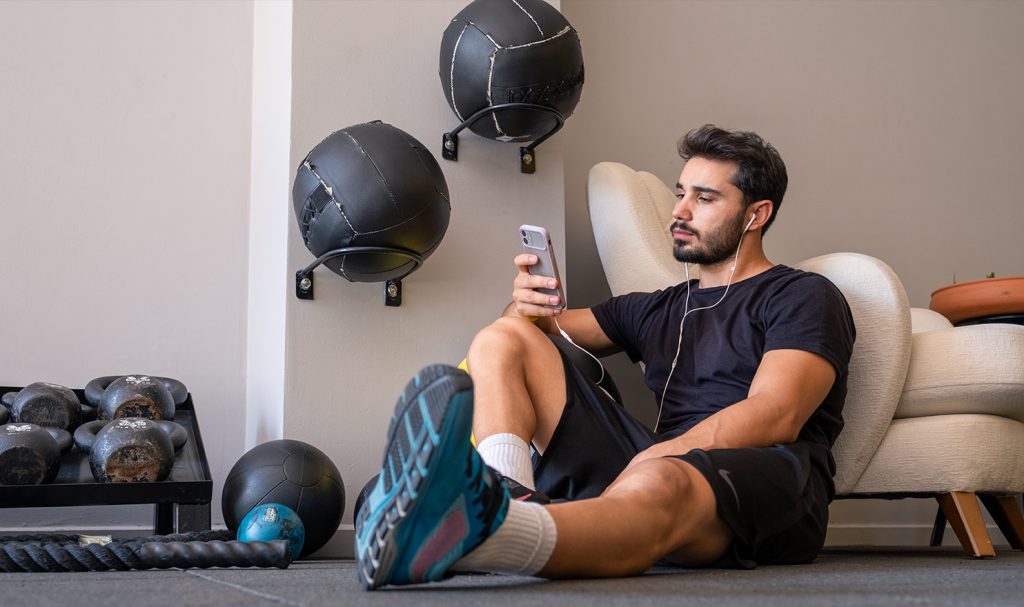 A young man sits on the floor of a fitness room in his home with his back resting against the side of a couch. He is reading his weekly curriculum on his phone for his personal trainer certification.