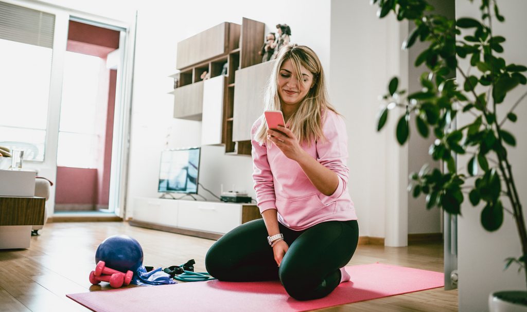 A woman kneels on the yoga mat in her home as she reads her daily studies for her personal trainer certification program on her smart phone.