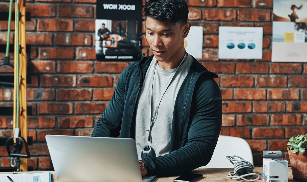 A young man sits at a desk in a fitness room while he works on his personal trainer certification program from his laptop.