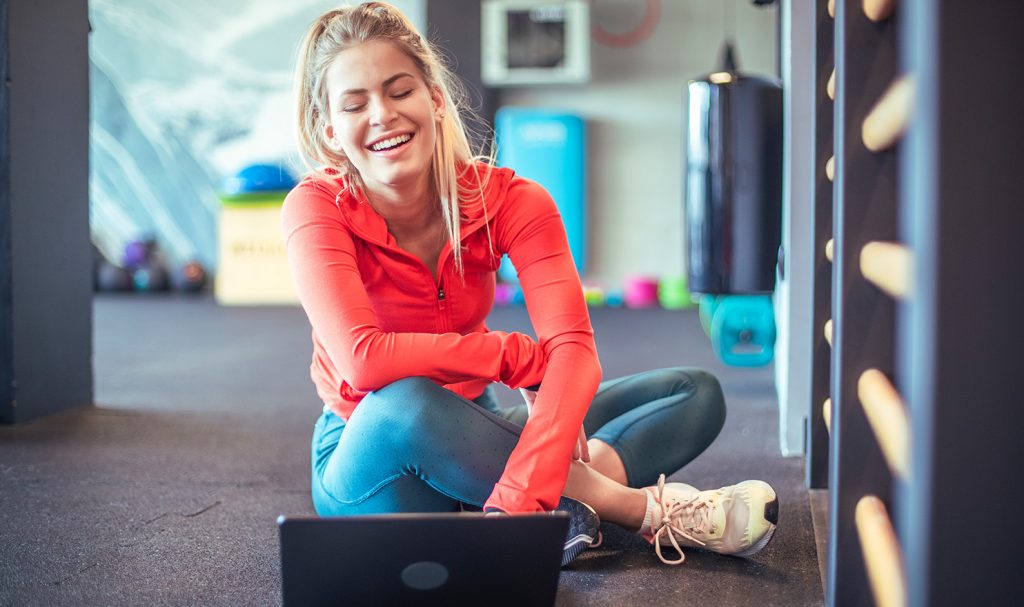 A woman sits on the floor of a fitness center as she happily works on her personal trainer certification on her laptop.