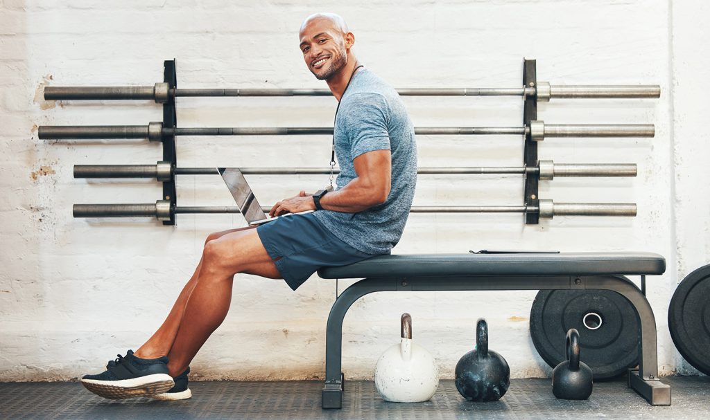 A man sits on the edge of a workout bench next to kettle weights in a workout room as he works on a laptop to receive his personal trainer certification.