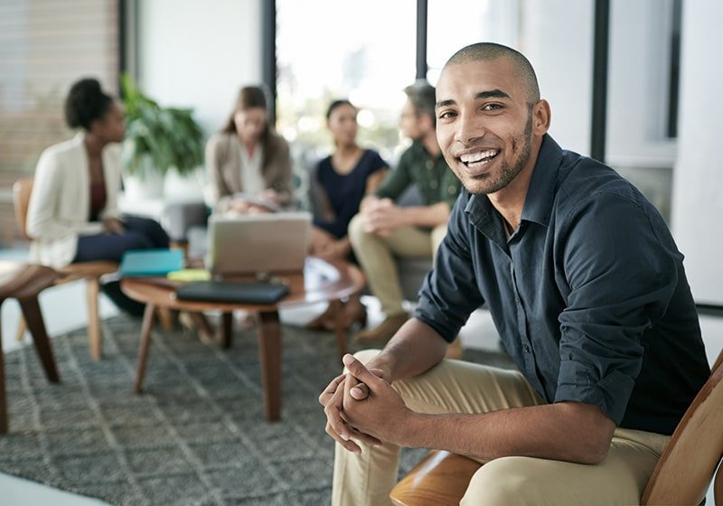 A team member smiles with the team on a couch in the background.