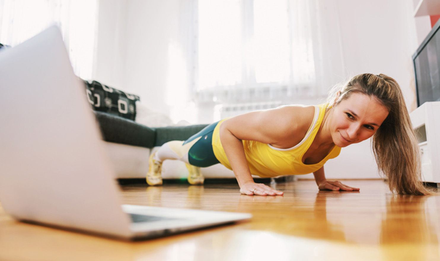 A virtual trainer is demonstrating a plank in front of her computer monitor on the floor of her home as she guides her client in an online workout.