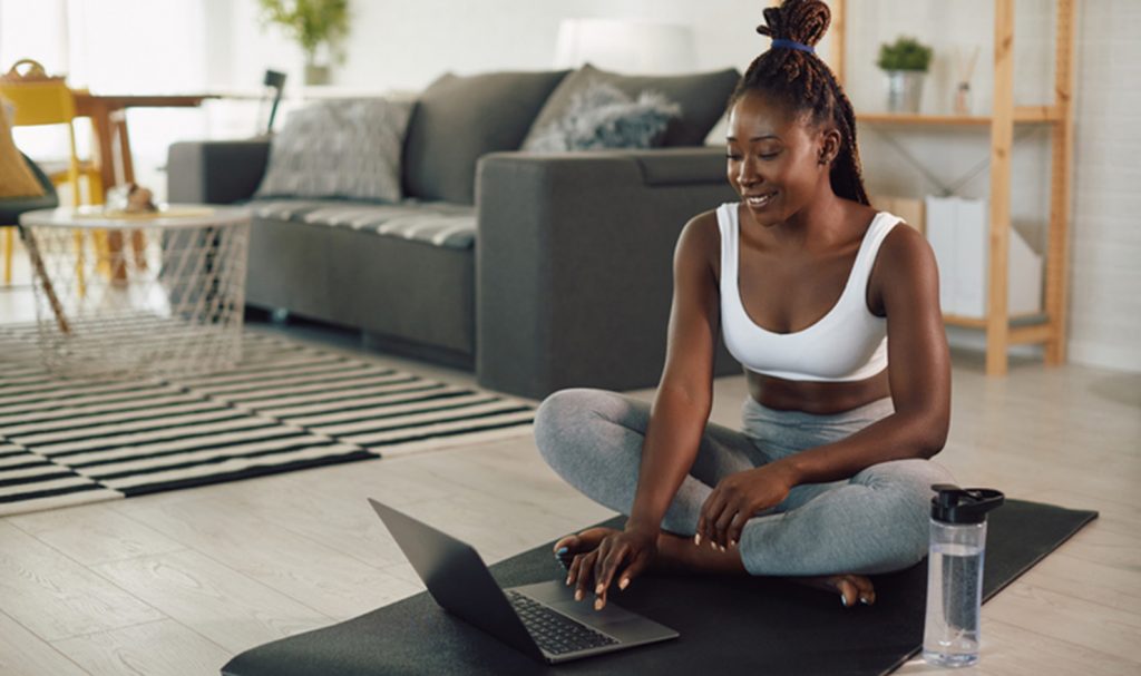 A young woman sits on a yoga mat in her living room as she smiles at her laptop monitor where she watches workout videos her trainer filmed for her.