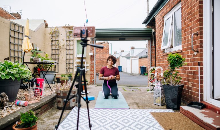 An online personal trainer is recording a session for social media outside of her city apartment on a patio. She is kneeling on a blue yoga mat and speaking to her camera on a tripod.