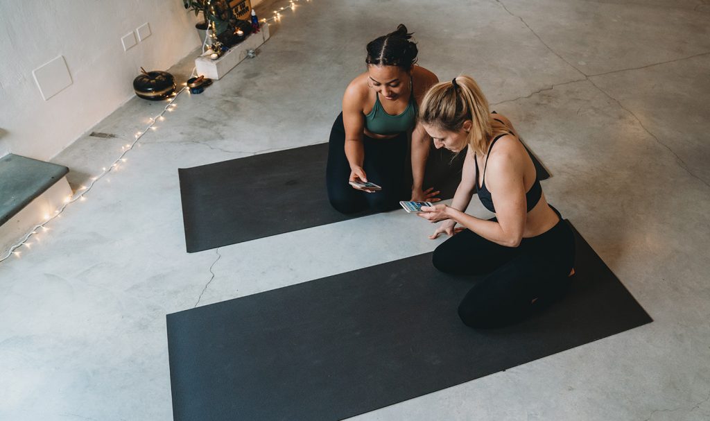 Two online yoga teachers sit on gray yoga mats on the ground inside a private studio. They are on their phones networking with each other to grow their online training businesses.