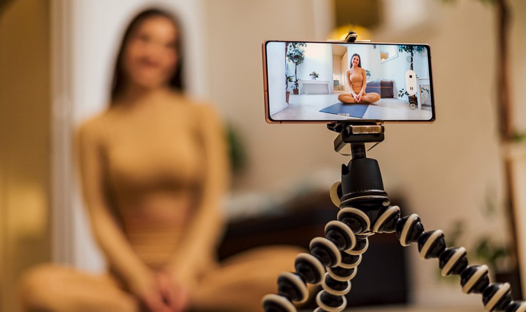 The image is focused on a smart phone on a tripod that is a filming a young woman in her home. She is sitting on a mat with weights as she captures video content for her social media pages to help her find new personal training clients.