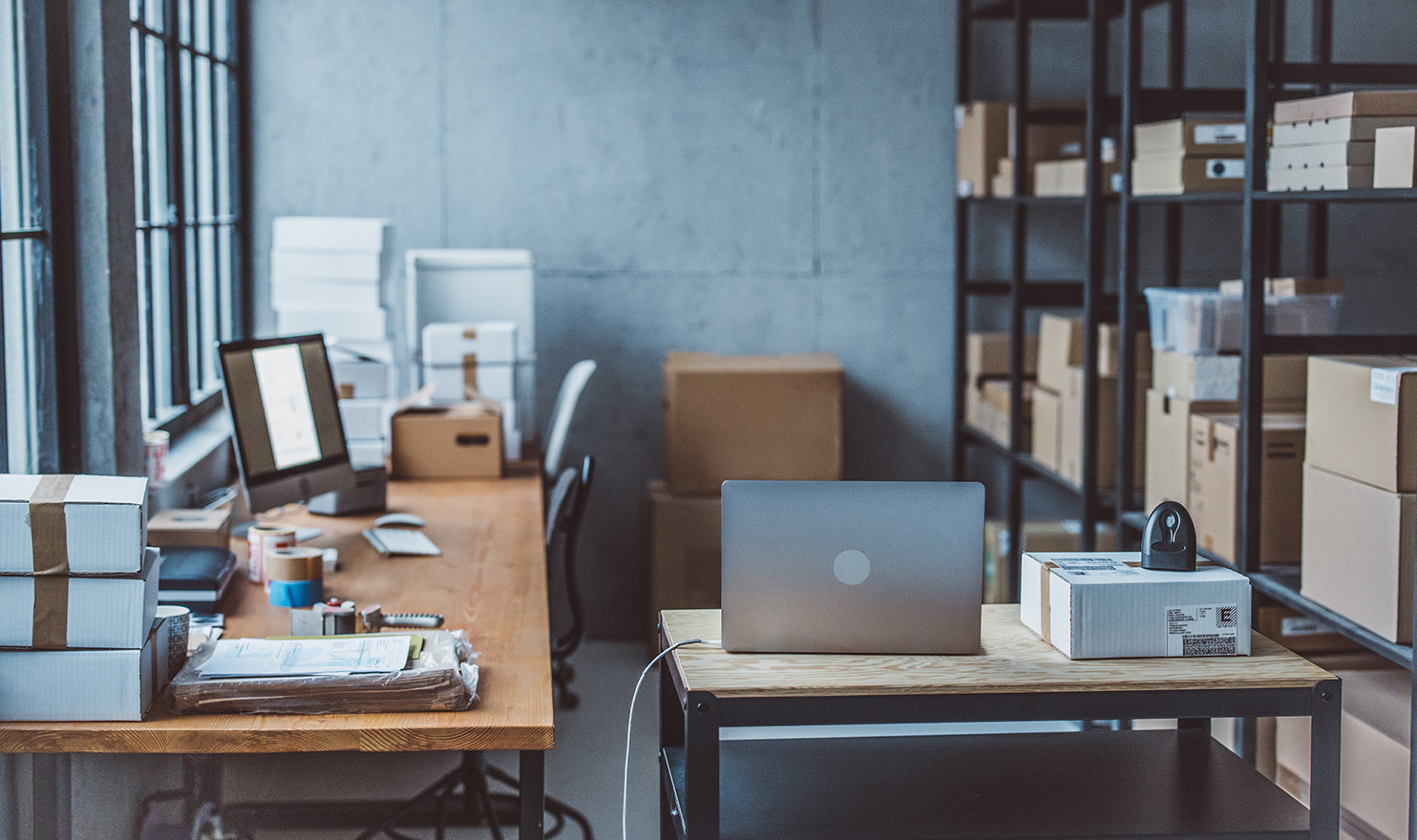 Two desks sit in a small business office surrounded by the supplies and inventory used to run the business and computers with software used to manage the business operations.