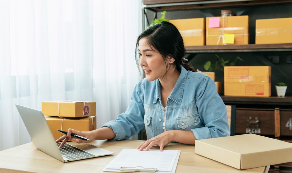 A young woman sits at a desk in her home office as she works on tracking her shipments and orders for her new business.