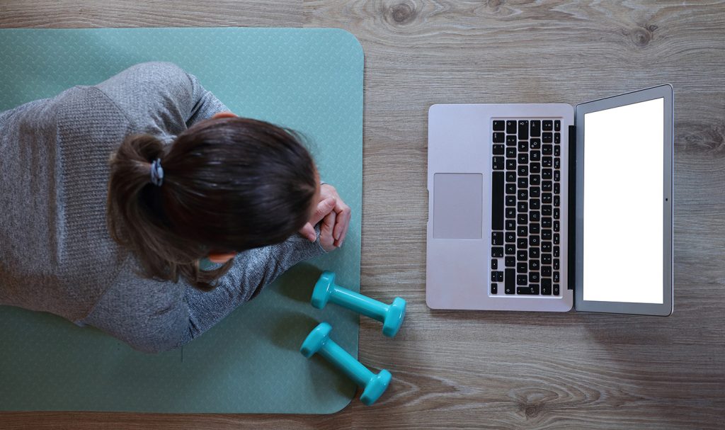 A virtual personal trainer lays on a yoga mat next to weights as she works on her laptop to schedule out her clients for the week.