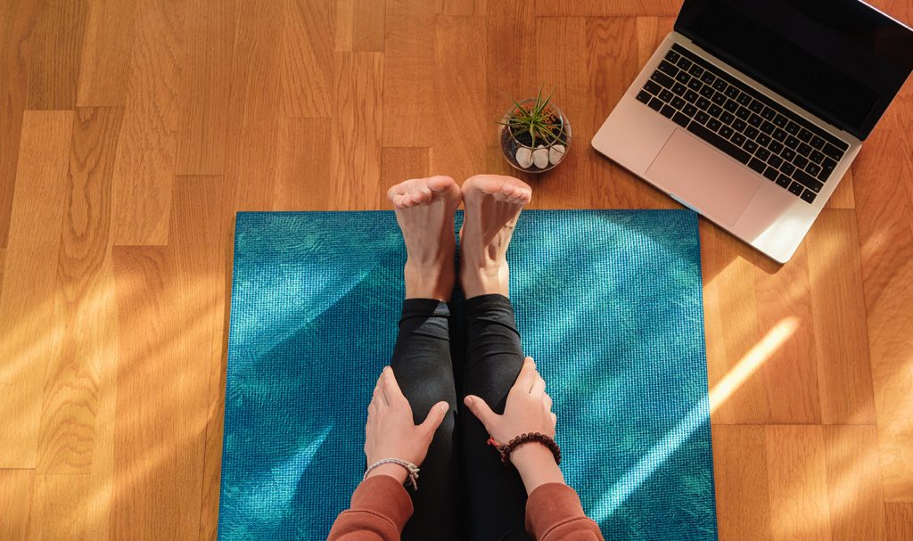 A pair of outstretched feet on a blue yoga mat are next to a small green succulent plant and a laptop were the virtual trainer prepares to call his next virtual client for the day.