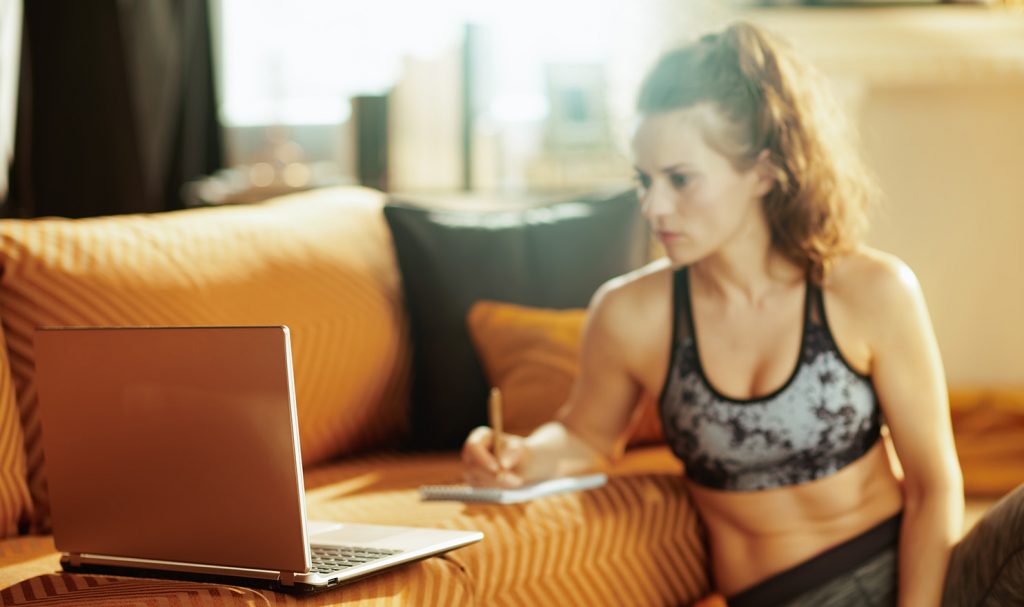 An online trainer is sitting on the ground of her home propped up against the side of her couch where she is looking at her client's information on her laptop and writing out notes to make a personalized training plan for them.