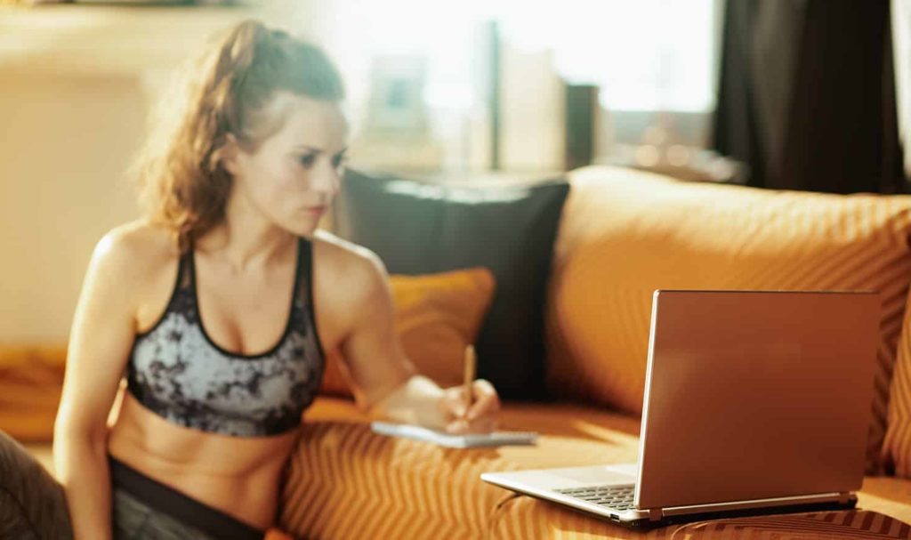 An online trainer is sitting on the ground of her home propped up against the side of her couch where she is looking at her client's information on her laptop and writing out notes to make a personalized training plan for them.
