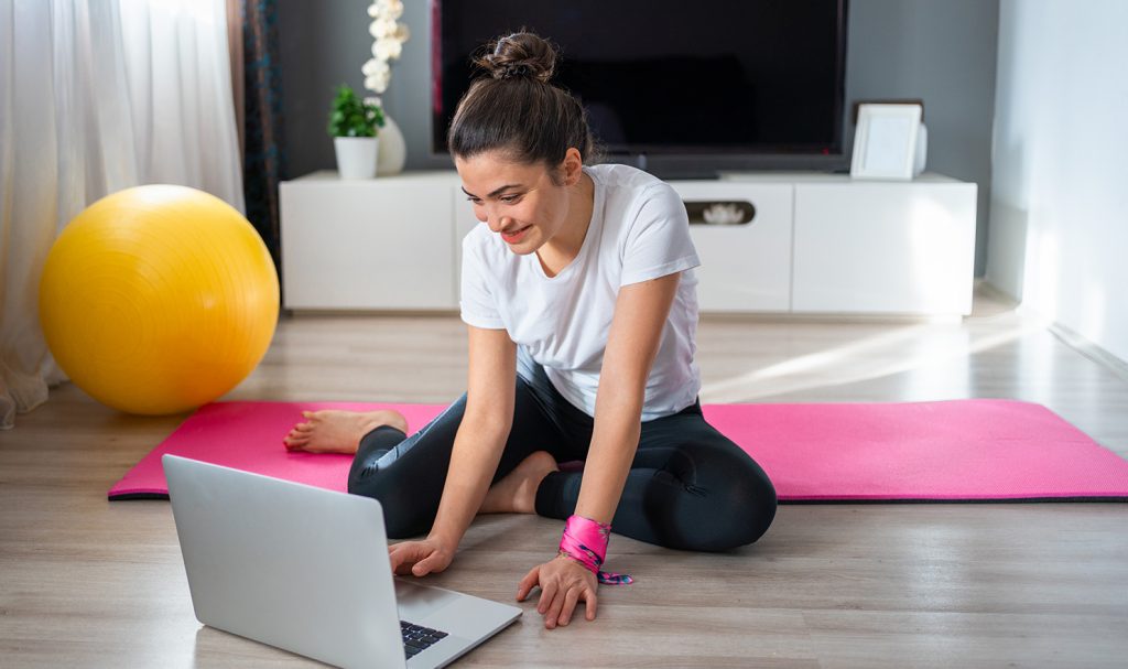 A virtual personal trainer is sitting on a pink workout mat in her living room as she works on organizing her client data on her computer to keep her more organized.