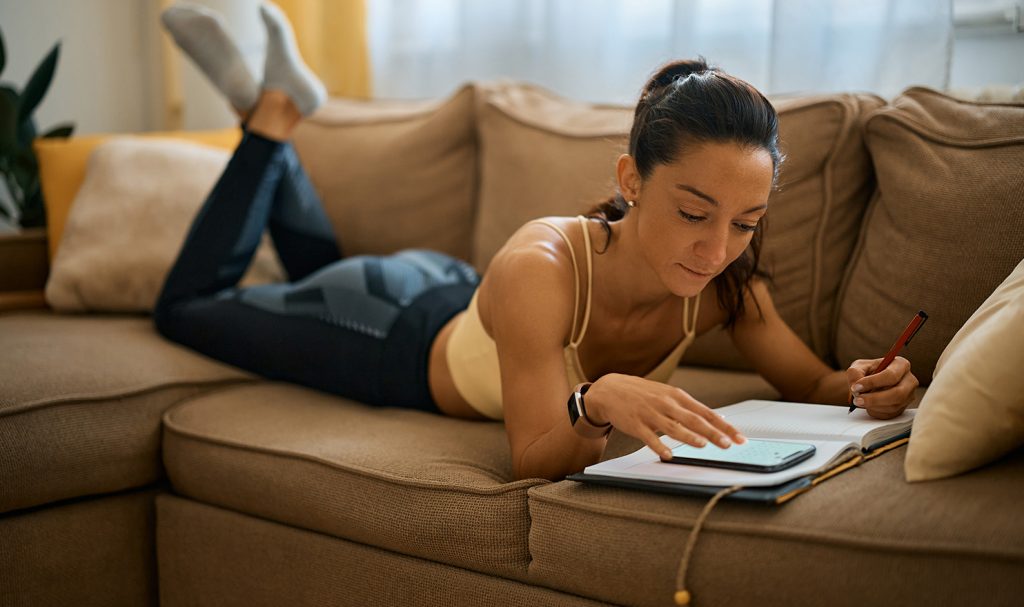 An online trainer is lying on her stomach on her couch as she checks her calendar in her notebook and prepare reminder text messages on her phone for her virtual clients.
