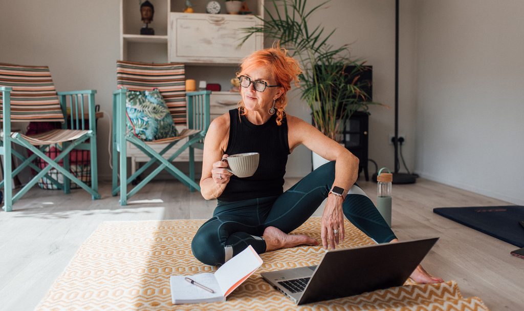 An online personal trainer is sitting on a workout mat in her living room while she works on her laptop sipping on coffee next to her planner with a calendar of her upcoming client trainings.