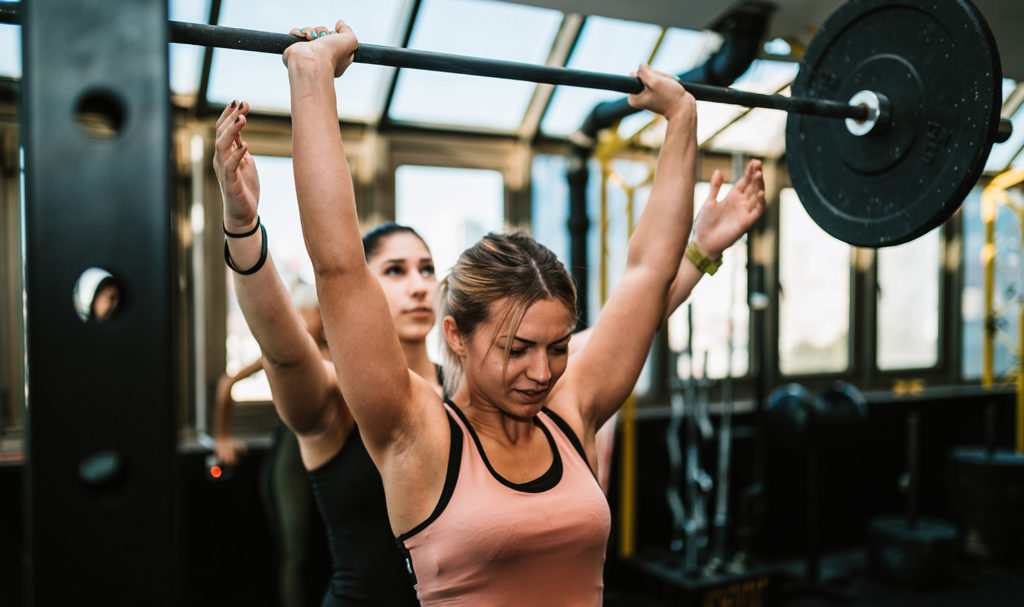 A female personal trainer is helping her female strength training client with her weight training by cautiously watching her hold a bar of weights over her head.