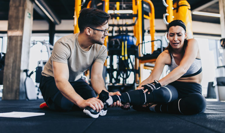 A trainer with personal trainer insurance is helping his client who is sitting on the ground in the gym after twisting her ankle during a workout session.