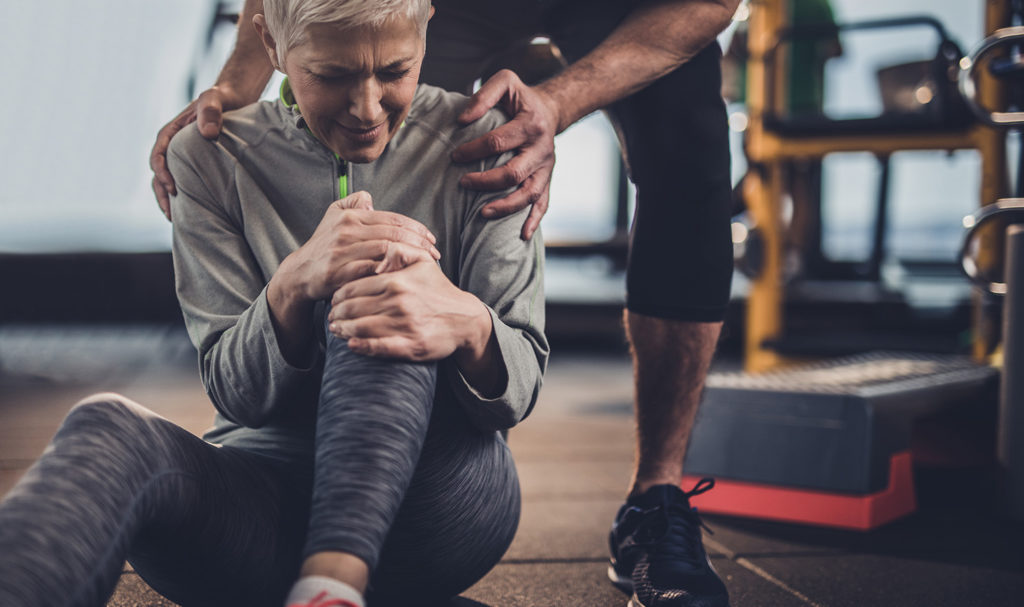 A women sits on the floor gripping her knee in pain after hurting herself during a training session. Her personal trainer has his hands on her shoulders to comfort and calm her, as he knows his personal trainer insurance can help him pay for accidental injuries.