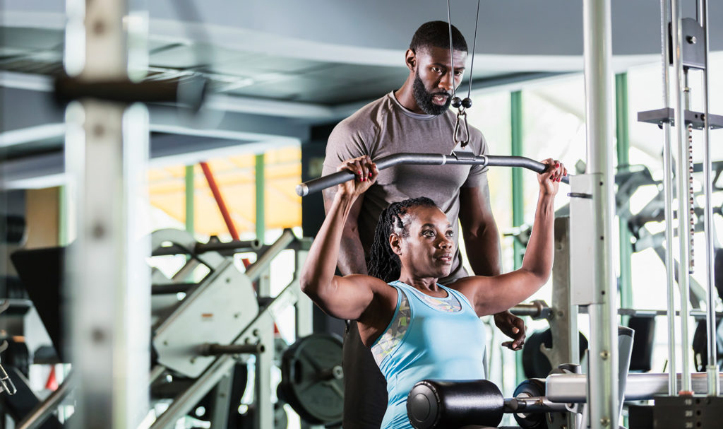 A personal trainer is standing behind his client as she is at a seated chest press machine. He carefully guides her movements and knows if she is hurt he has personal trainer insurance to help him.