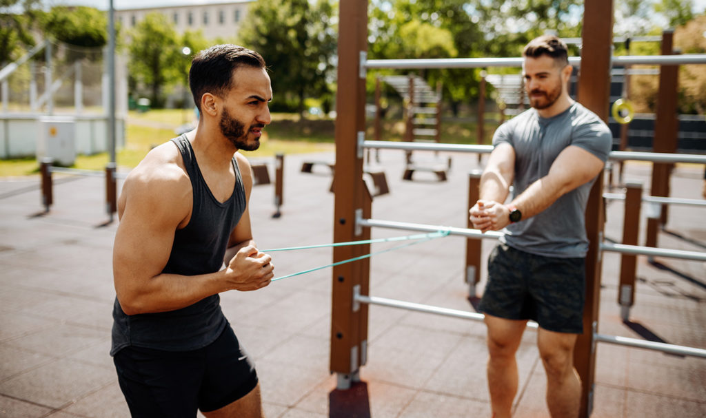 A private trainer with personal trainer insurance is showing his client the right way to hold the resistance band in his fists as he trains in a outdoor workout space.