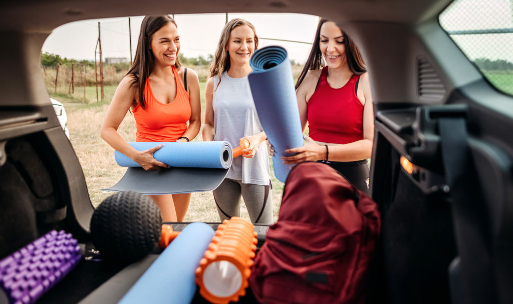 Having personal trainer insurance makes transporting equipment easier for this trainer. Her and two clients are unloading workout gear from the trunk of the car together for their personal training session.