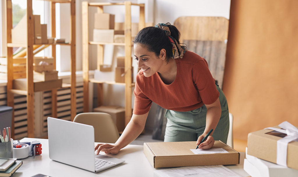 A small business owner insured with product liability insurance is bending down to look at her laptop on her desk while preparing a label for a package. She is mailing out items from her small online shop out of her home office where she is surrounded by shelves of products and boxes.