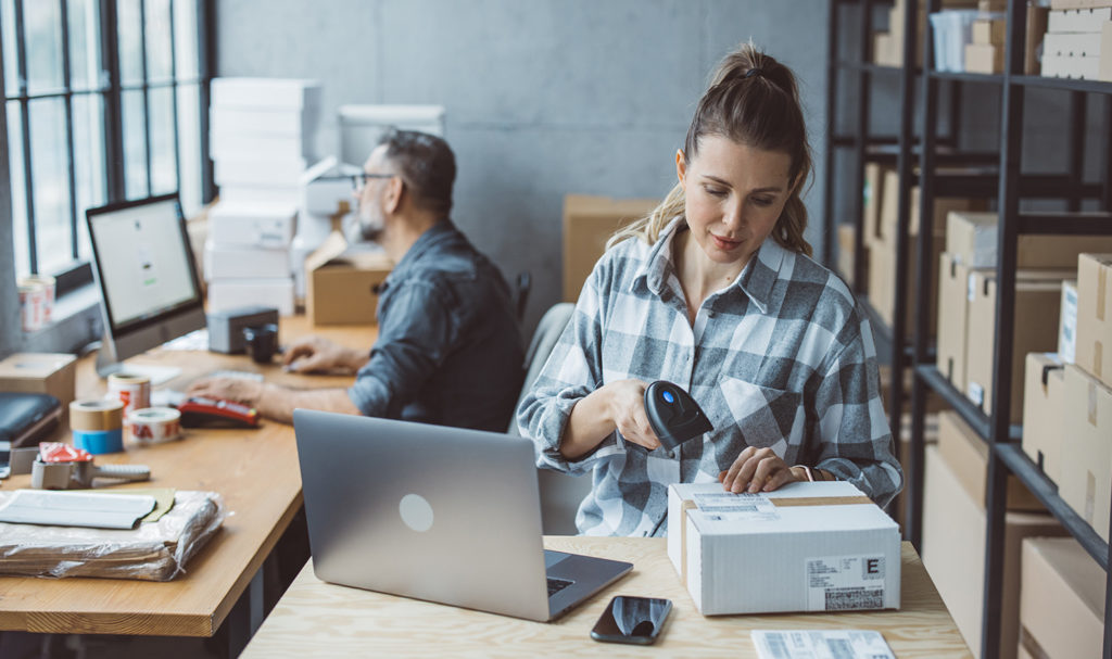 A boss and his employee are working on packaging items for shipment in their office space with shelves full of product and packages.