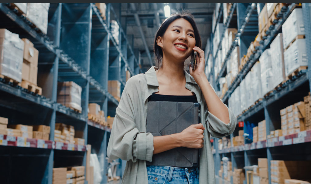 A young business owner is smiling on the phone while walking through an aisle of tall shelving in a warehouse. The business owner is clutching a clipboard where they are listing out all of their products for their product liability insurance policy.