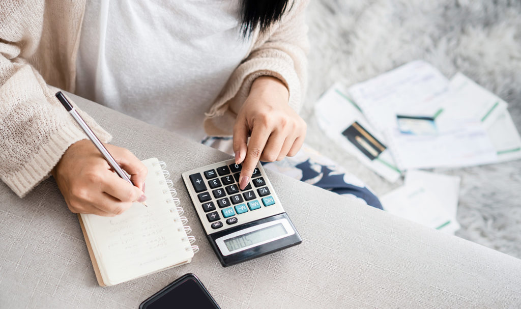 A woman using a calculator is punching in numbers while taking notes on a notepad and a pile of papers sit next to her. She is calculating the cost of her product liability insurance.