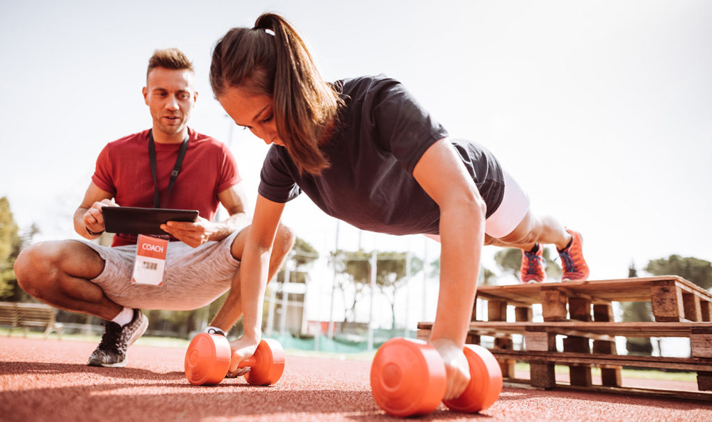 A young woman is doing push ups on a set of weights as her trainer kneels to the side of her with a clipboard guiding her through a workout.