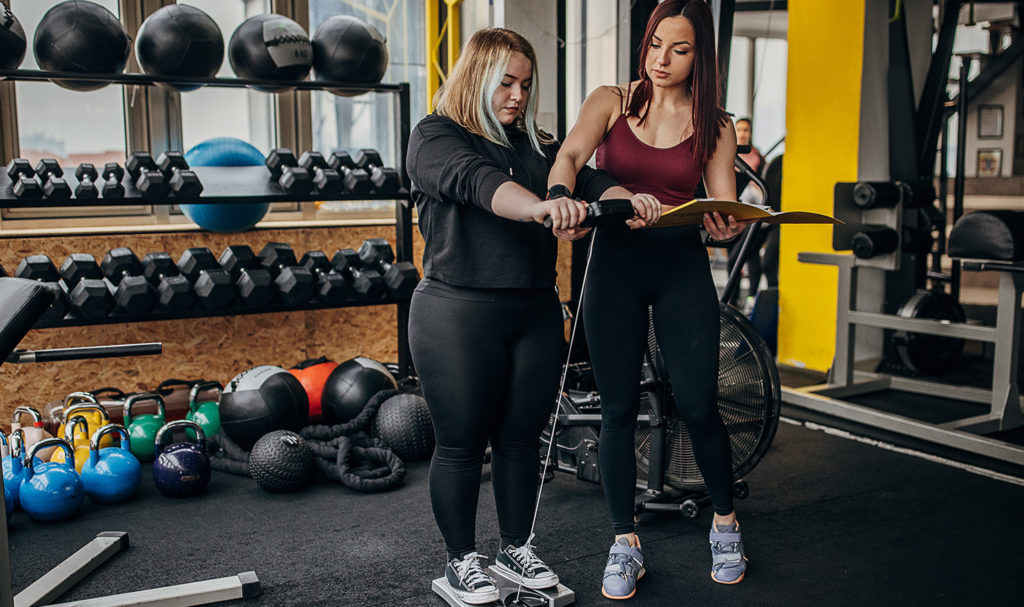 A personal trainer guides her client through how to use a standing resistance machine as part of her workout routine.