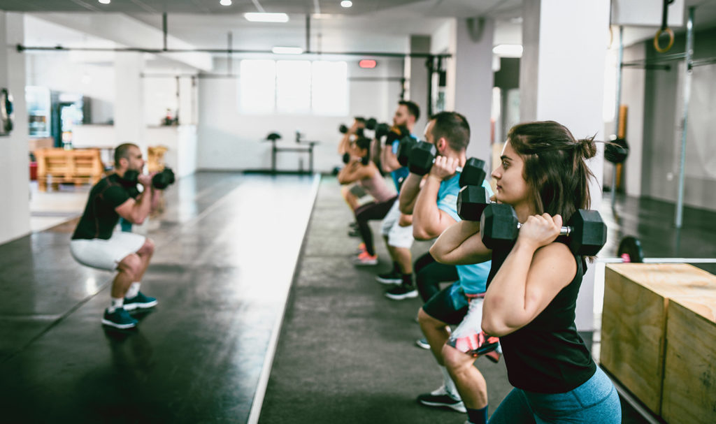 A group of students in a fitness class are doing squats with weight in their hands as they follow the instruction of their teacher at the front of the class.