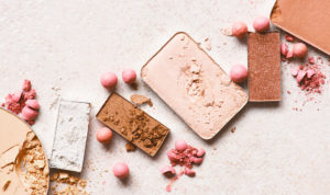 A set of broken makeup palettes lay on a tan background. Product liability insurance may be able to cover damages to beauty products like these eye shadow sets.