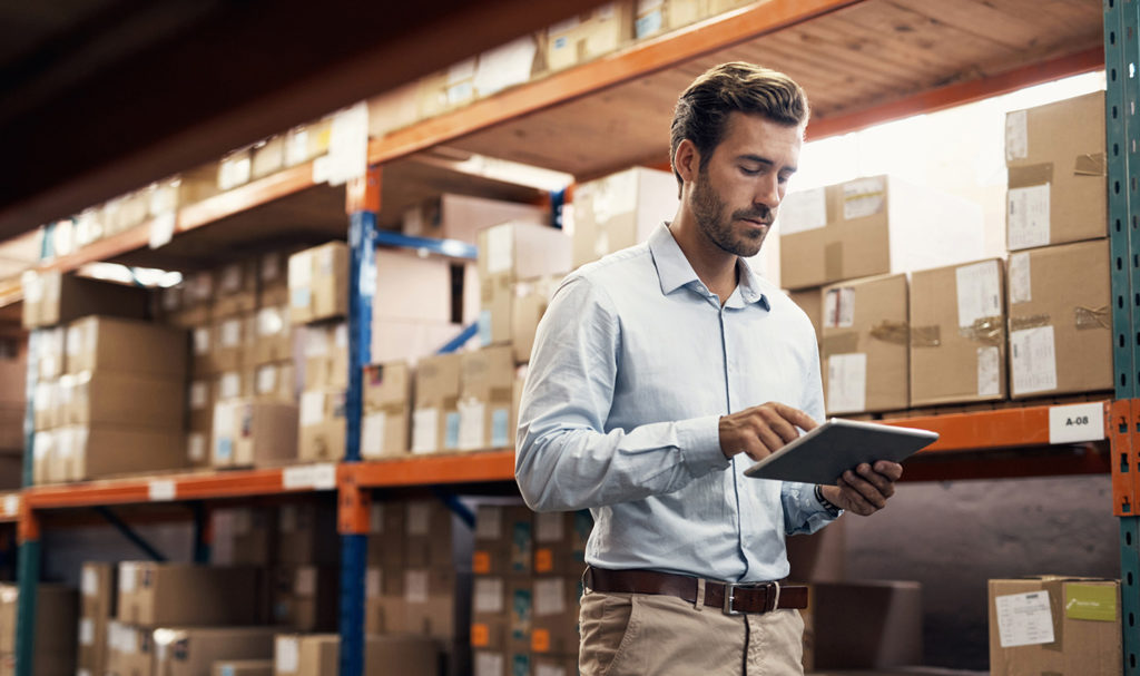 A young man in a dress suit and slacks is walking through the aisles of a warehouse where boxes of unsold products sit on a shelf. He is taking inventory on a notepad for his product liability insurance policy.
