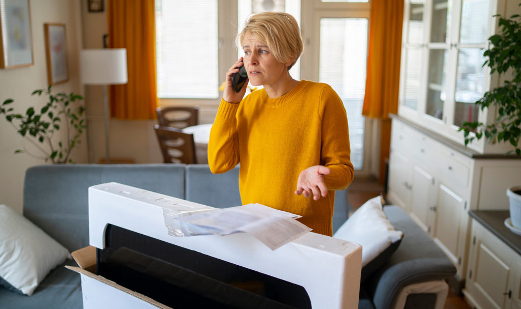 A woman is visibly upset as she sits on the phone with a customer service rep because a product she ordered was broken and damaged in the package. This type of damage may be covered with product liability insurance.