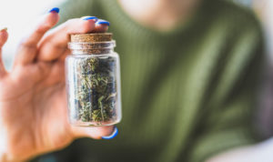 A woman in a green shirt sits out of focus holds a bottle of cannabis buds in a small glass vile up to the camera to be in focus.
